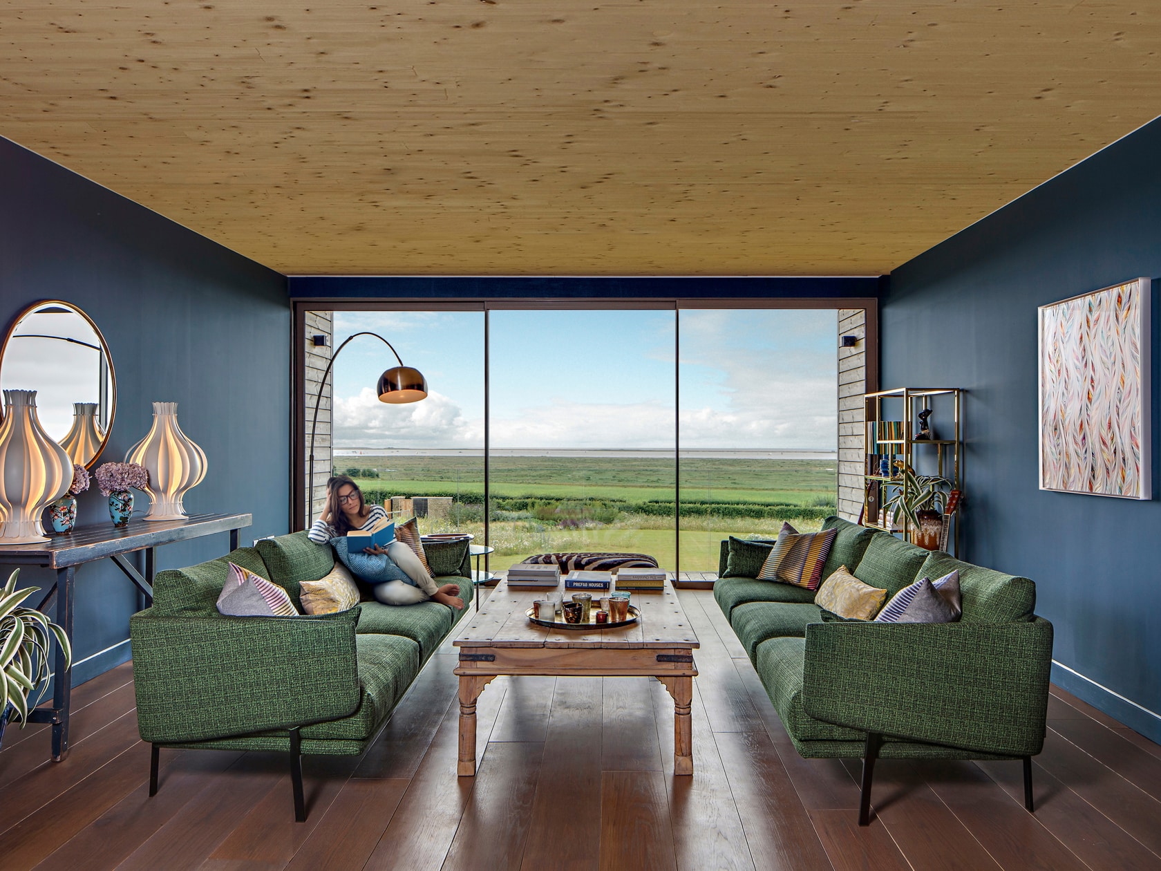 Luxury home living space featuring large glass windows, wooden panelling, green sofas and an outdoor patio with natural lighting.