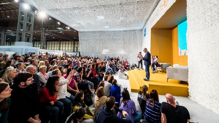 A speaker addresses a seated audience at an indoor event with bright lighting and textured white decor on the walls and ceiling.