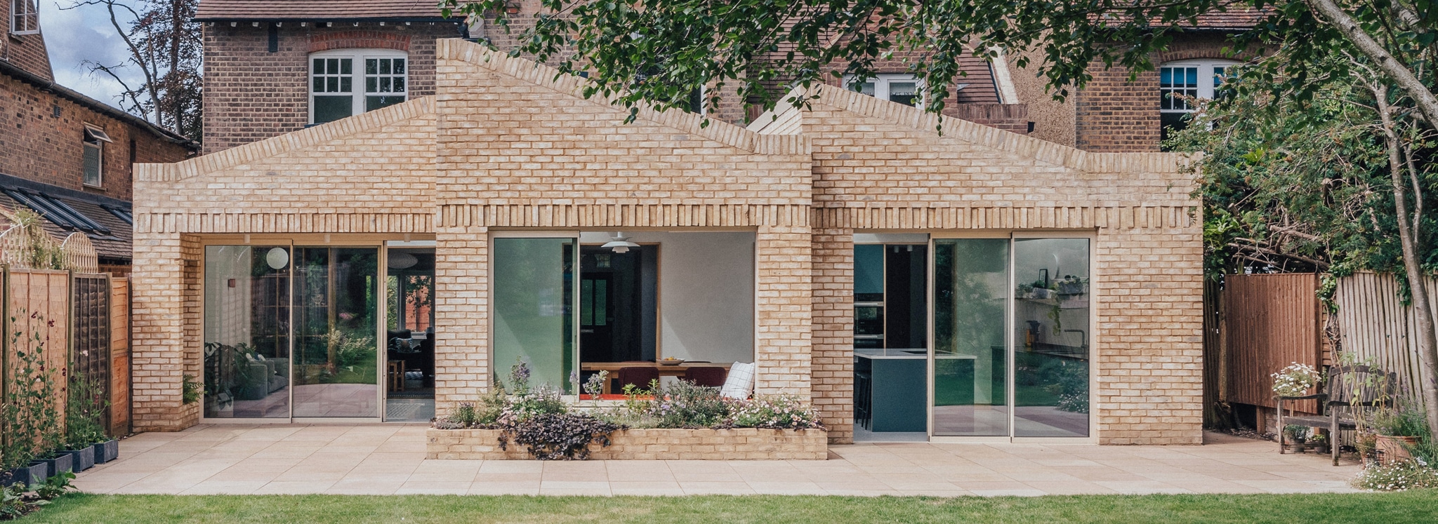 Single-story brick extension with pitched roofs, large glass doors, and windows, attached to a traditional brick house, overlooking a paved patio and garden.