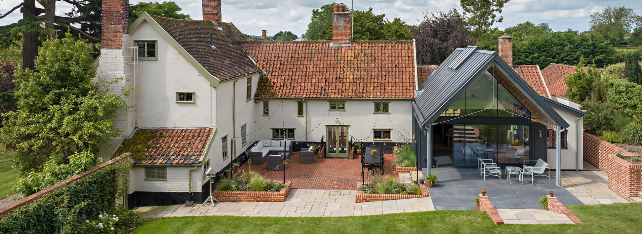 A large house with a red-tiled roof and modern glass extension, featuring outdoor seating areas, brick patio, and a landscaped garden.