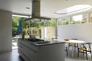 Modern kitchen with a central island, built-in stove, dining table with chairs, large windows, and a view of a green backyard. Natural light fills the space.