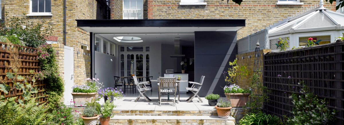 Modern patio with outdoor seating and potted plants leads to an open-plan kitchen extension at the back of a brick house.