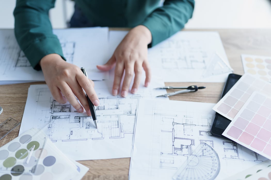 Person reviewing building floor plans with architectural drawings, color swatches, and drafting tools spread out on a wooden desk.