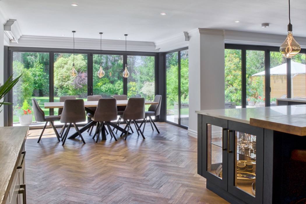 Modern dining area with large windows, a wooden table and grey chairs, overlooking a lush garden; kitchen island with marble countertop in the foreground.