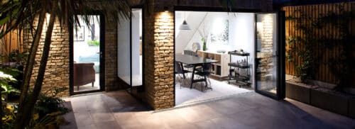 A modern dining room with brick exterior walls opens onto a tiled patio through large glass sliding doors; indoor and outdoor spaces are illuminated at night.
