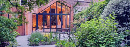 A modern glass-walled extension with wood siding opens onto a lush, green garden with various plants and a stone patio path.