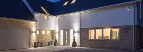 Modern house exterior at dusk with brick and white walls, large windows, illuminated entryway, and attached garage. Warm indoor lighting is visible through the windows.