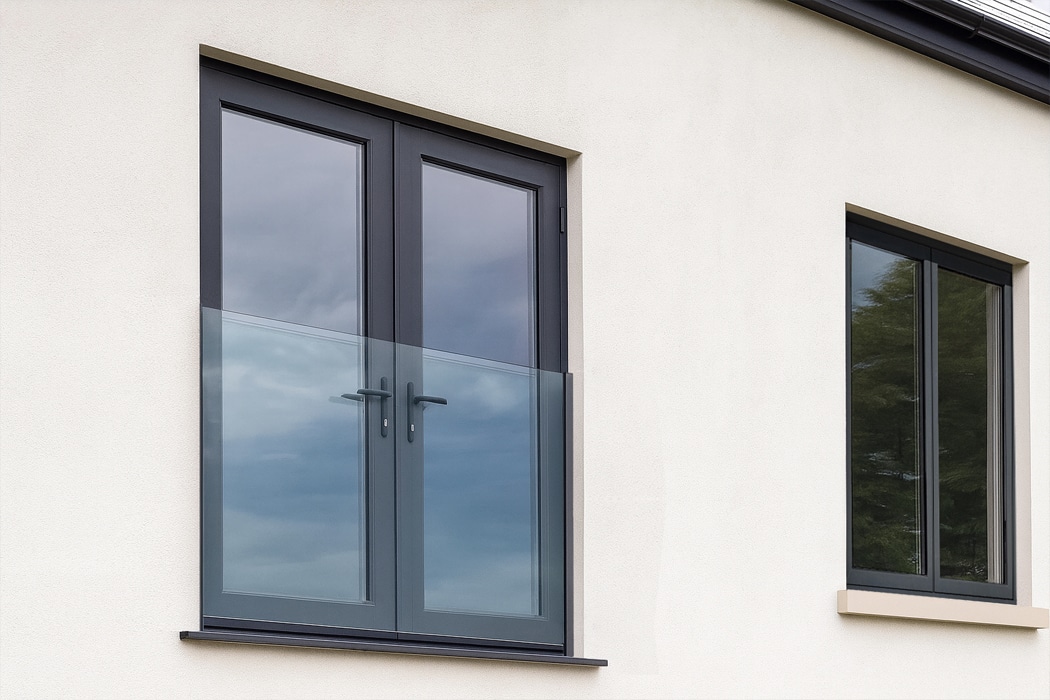 Modern grey aluminium French doors with slim frames and glazed panels, reflecting sky and trees against white rendered walls.