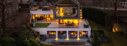 Modern multi-story house with large windows, outdoor lighting, and spacious terraces surrounded by greenery in the evening.