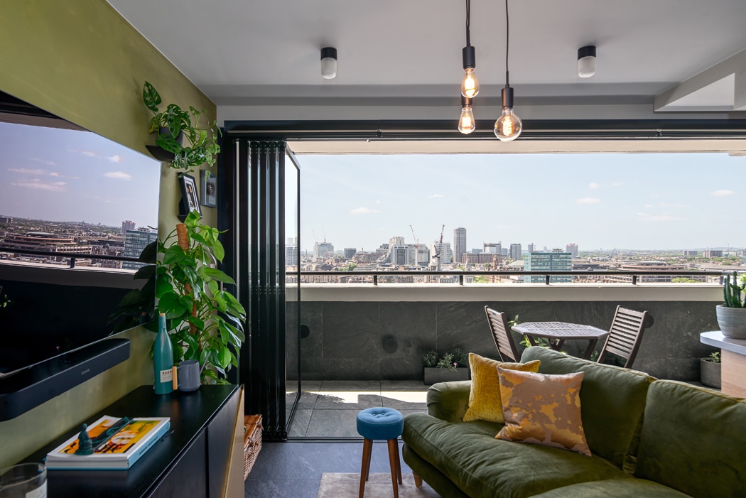 Modern living room with green sofa and indoor plants, opening to a balcony with a city skyline view under a clear sky. Round table and chairs are visible on the balcony.