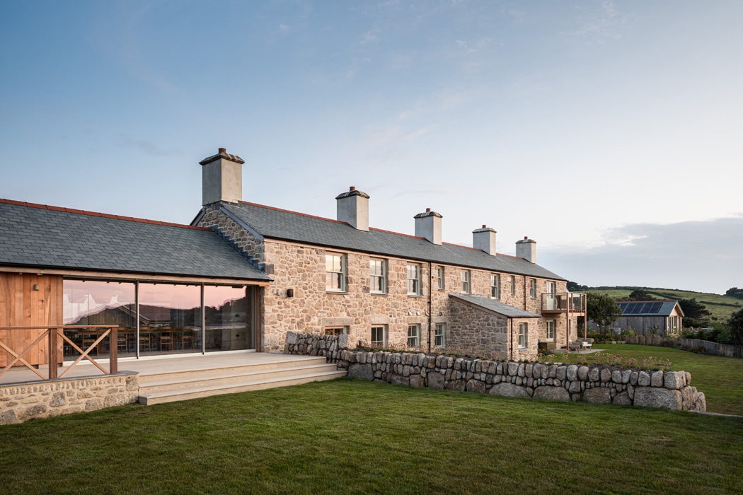 A row of stone cottages with chimneys and a slate roof stands behind a stone wall on a grassy lawn, set against a rural landscape at sunset.
