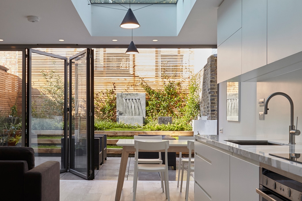Modern kitchen and dining area with sliding glass doors open to a patio and small garden. Natural light enters through a skylight and large windows.