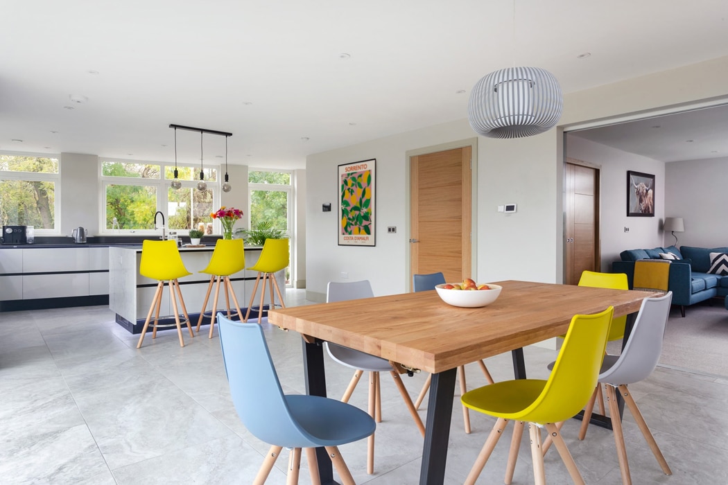 Modern open-plan kitchen and dining space featuring wooden table with coloured chairs and natural light from large windows.