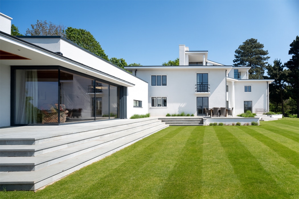 Modern white two-story house with large windows, flat roof, wide steps, and a neatly mowed green lawn under a clear blue sky.