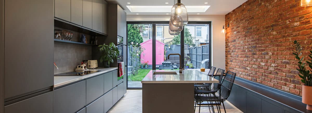 Modern kitchen with gray cabinets, an island with bar stools, exposed brick wall, large windows, and a view of a backyard with a pink shed and green lawn.