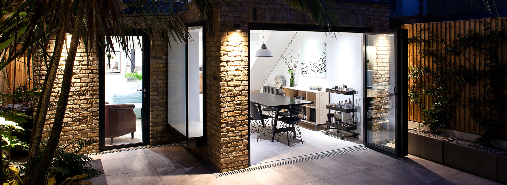 Modern dining area with glass doors, exposed brick walls, and outdoor patio, featuring contemporary furniture and soft indoor lighting.