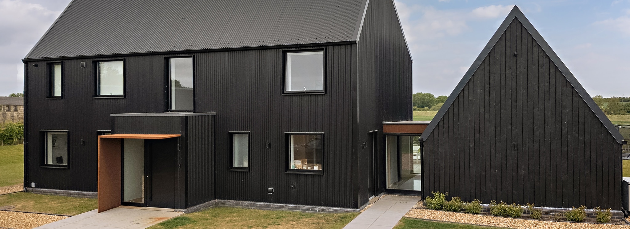 A modern black house with large windows, a sloped roof, and an attached angular extension, set in a grassy landscape under a partly cloudy sky.