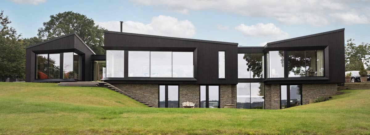 A modern two-story house with dark exterior panels, large glass windows, and stone accents, set on a grassy lawn with an overcast sky.