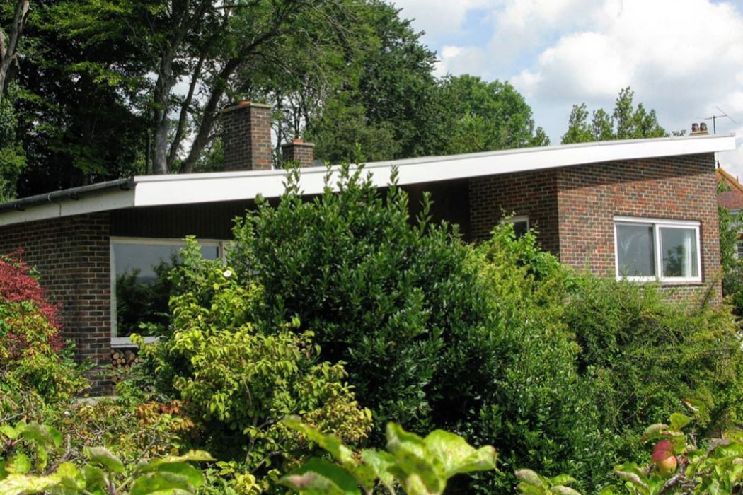 The original house on the plot, A brick house with large windows and a flat roof is partially hidden by dense green bushes and trees under a partly cloudy sky.