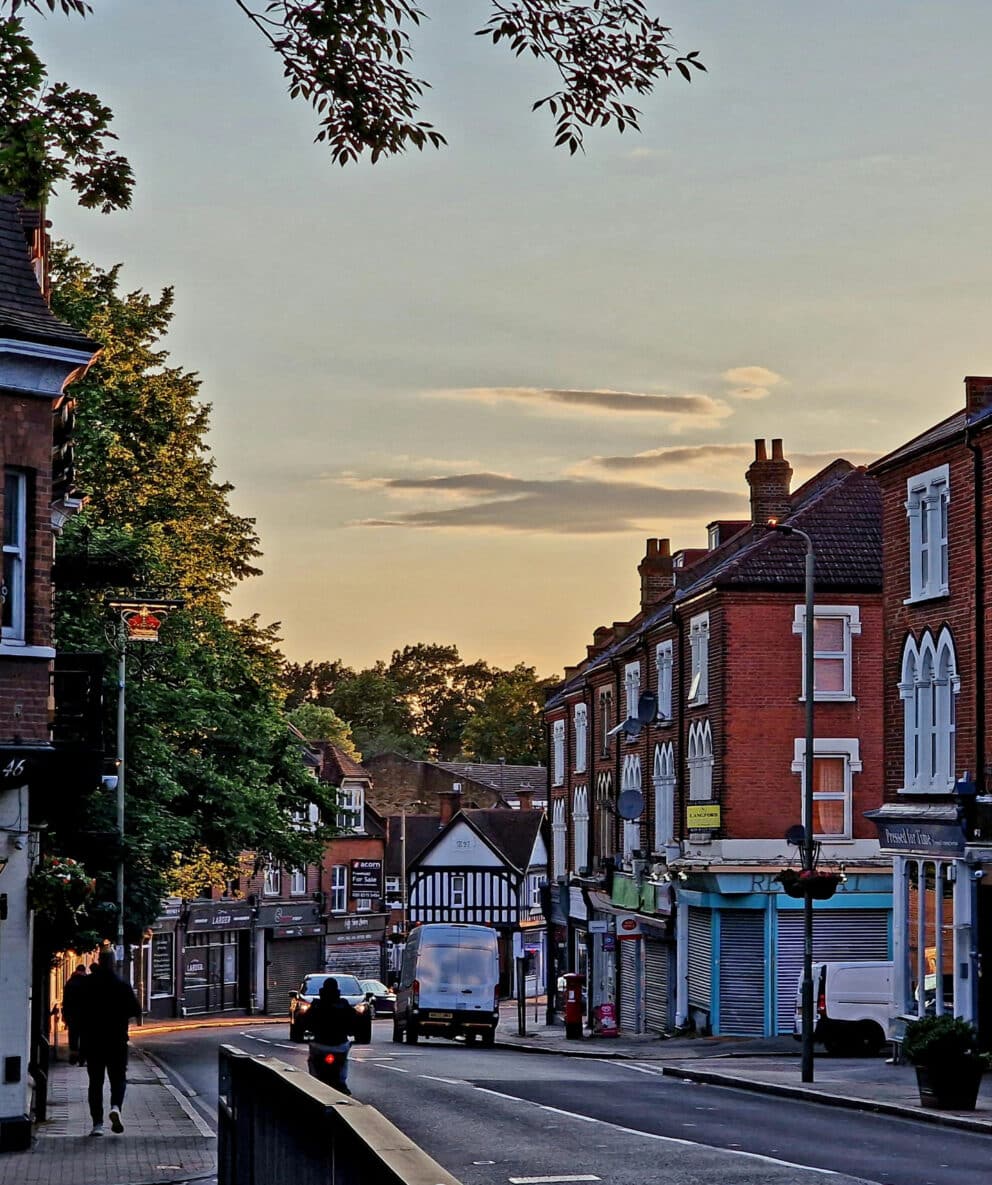 A quiet street scene at sunset with brick buildings, a few people walking, parked cars, and some shops with closed shutters.