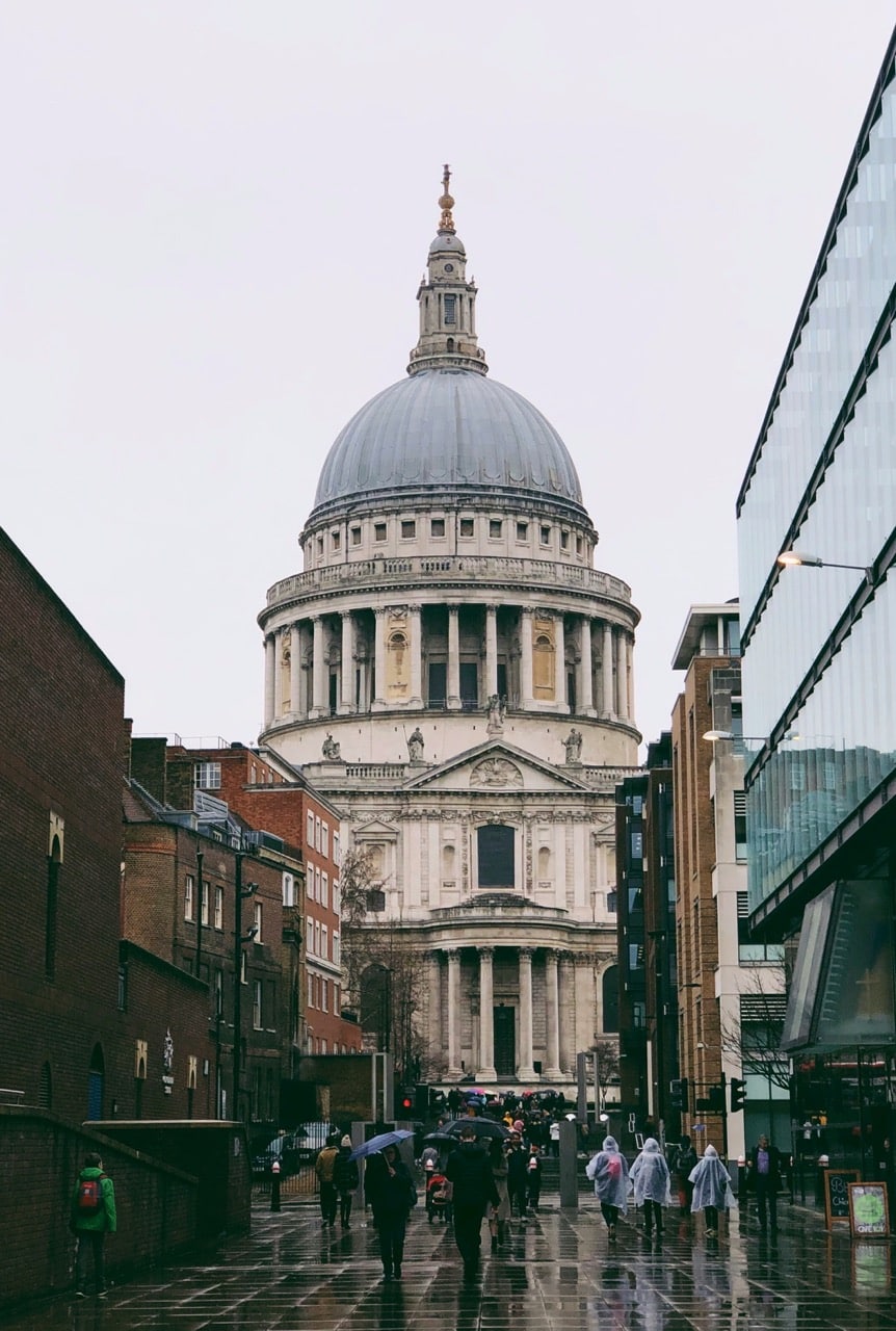 People walk along a wet city street towards St Paul’s Cathedral in London on a cloudy day, with modern and historic buildings lining the road.