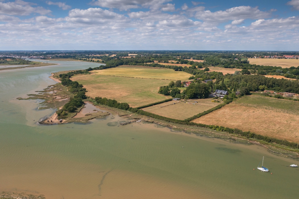 Aerial view of a contemporary riverside home in Essex with expansive fields and moored boats on the water below.
