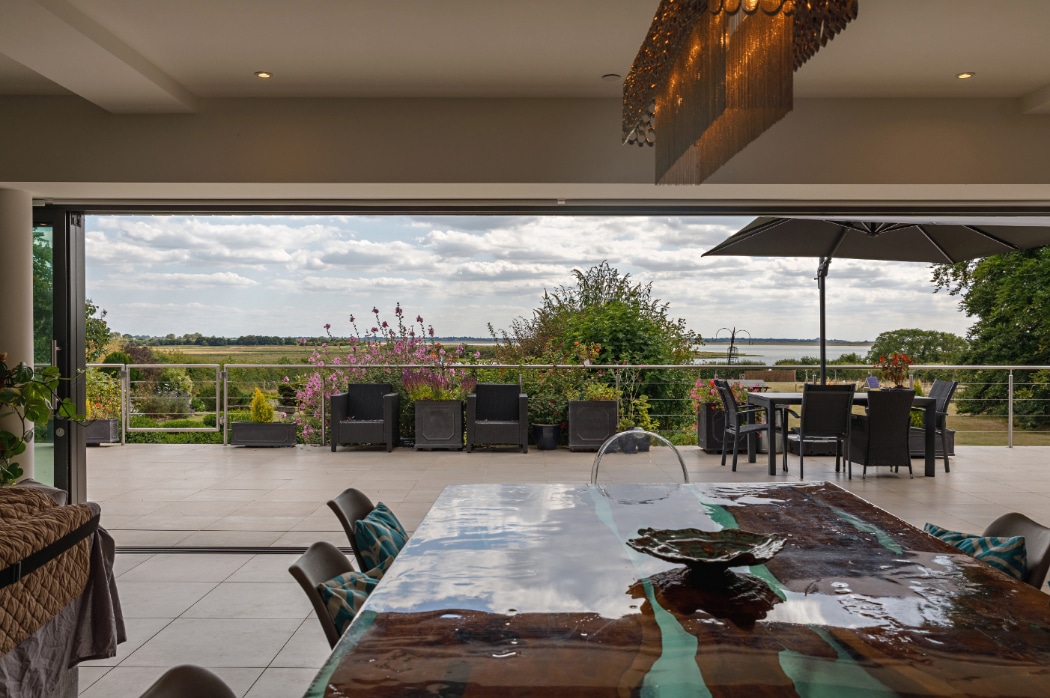Four panel sliding glass doors open onto a sun-lit riverside terrace overlooking Essex wetlands, seamlessly connecting modern interior to outdoor living space.