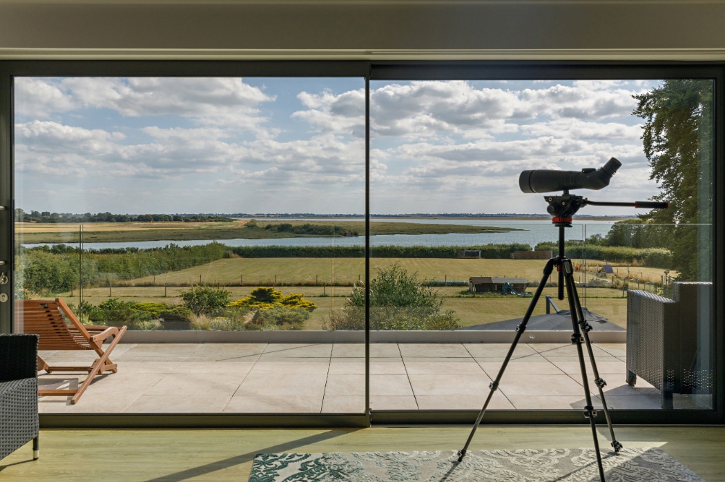 Contemporary riverside Essex home with 4 panel sliding glass doors opening onto a terrace overlooking fields and water.