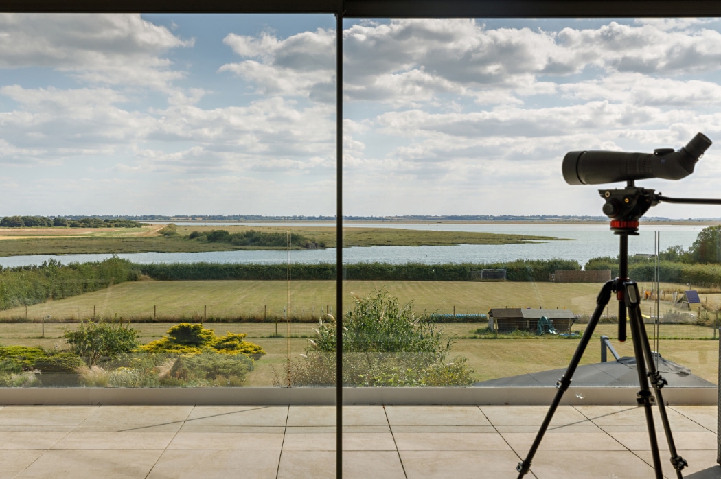 Spotting scope overlooking a contemporary riverside home in Essex with expansive views of water, fields, and natural landscape.