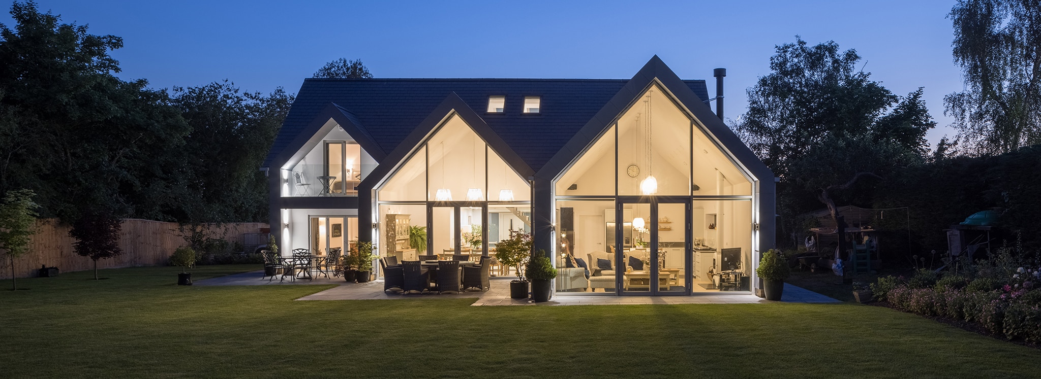 Contemporary house with expansive aluminium bifold doors and large glazed gable windows flooding the interior with light, set against an evening garden backdrop.
