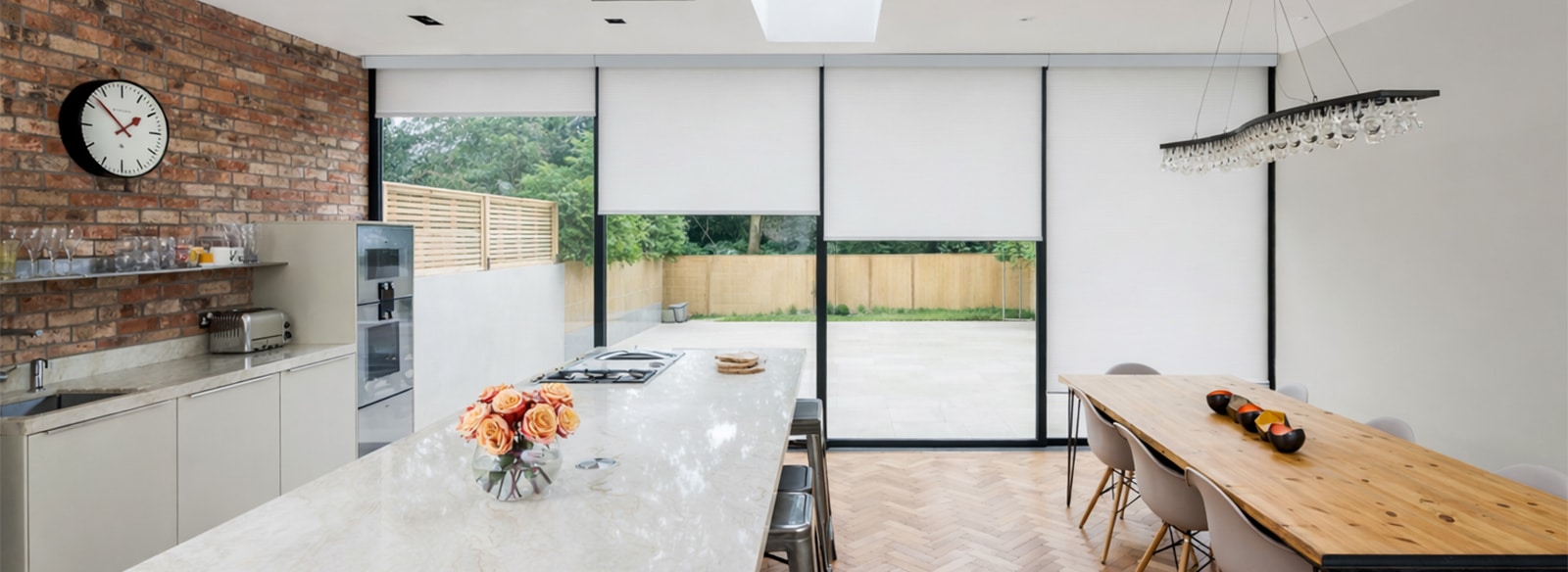 Modern kitchen with white roller blinds on expansive sliding doors overlooking garden, featuring marble island and wooden dining table.