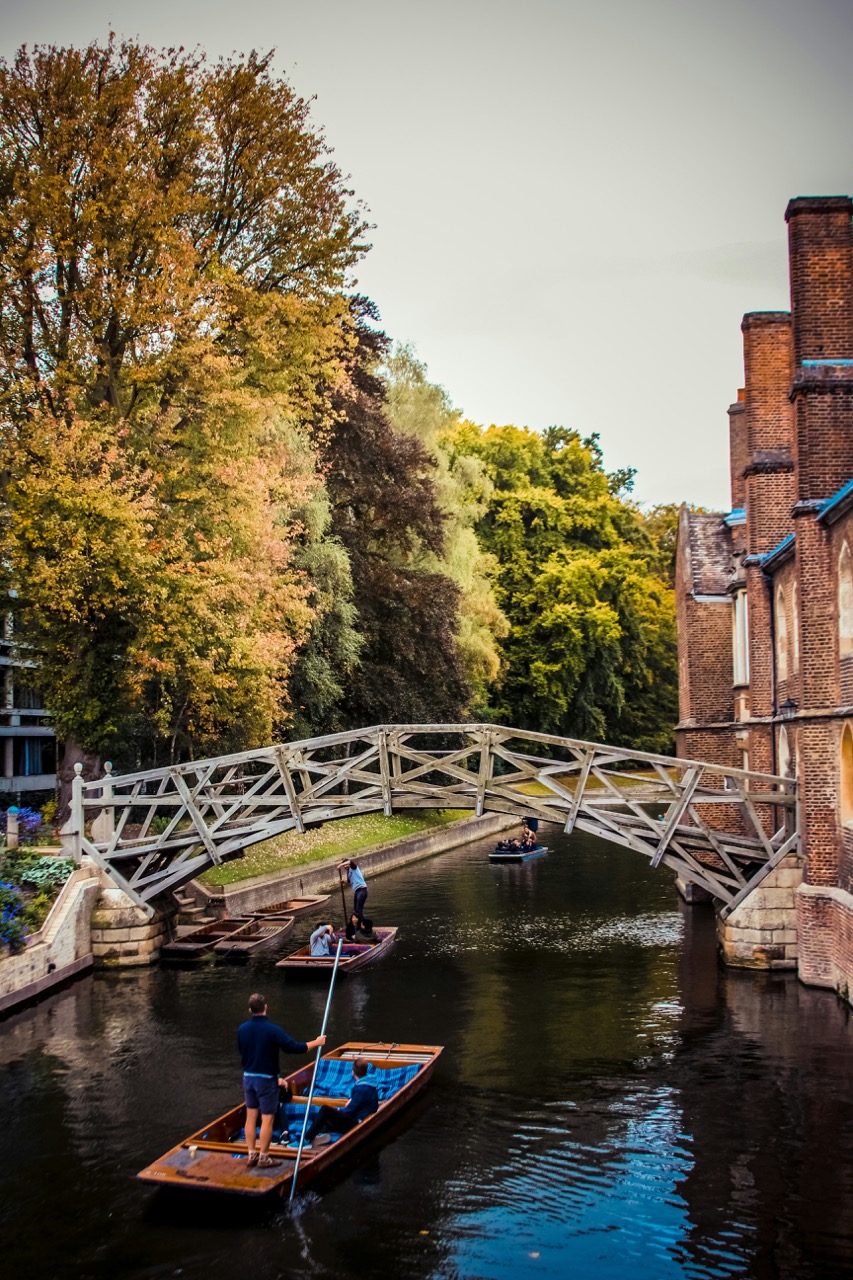 Punters navigate a serene canal beneath a wooden footbridge, framed by autumn trees and historic brick buildings in Cambridge.