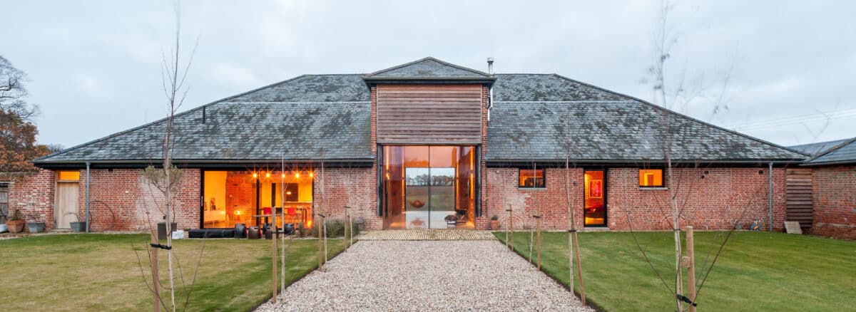 Contemporary barn conversion in Suffolk with expansive glazing, warm interior lighting, pitched roof, and gravel approach bordered by young trees.