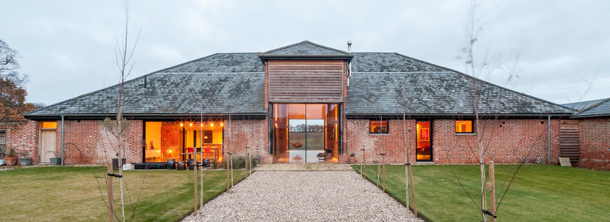 Contemporary barn conversion in Suffolk with expansive glazing, warm interior lighting, pitched roof, and gravel approach bordered by young trees.