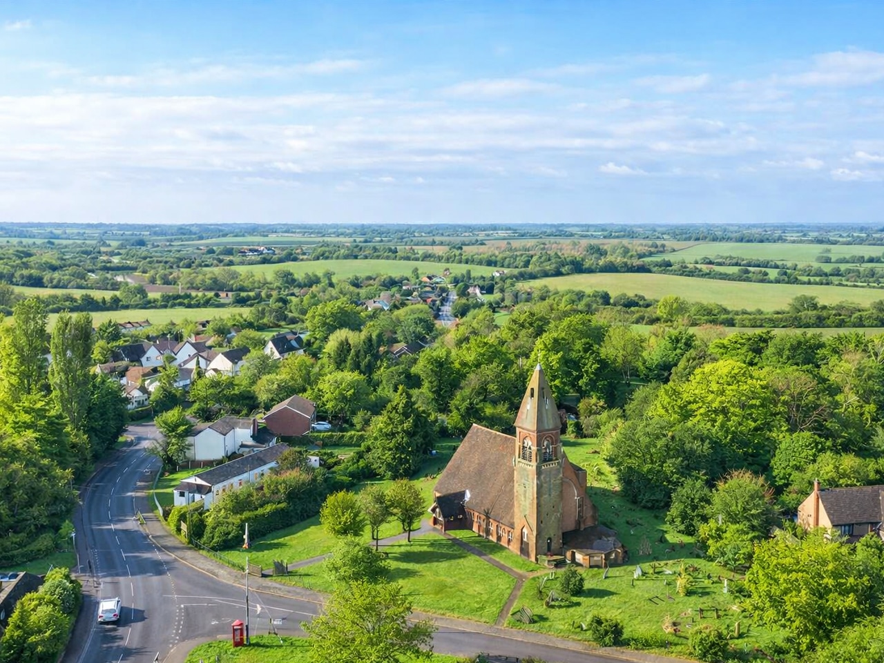 Aerial view of a charming English village with a brick church nestled amongst lush green trees and countryside fields.