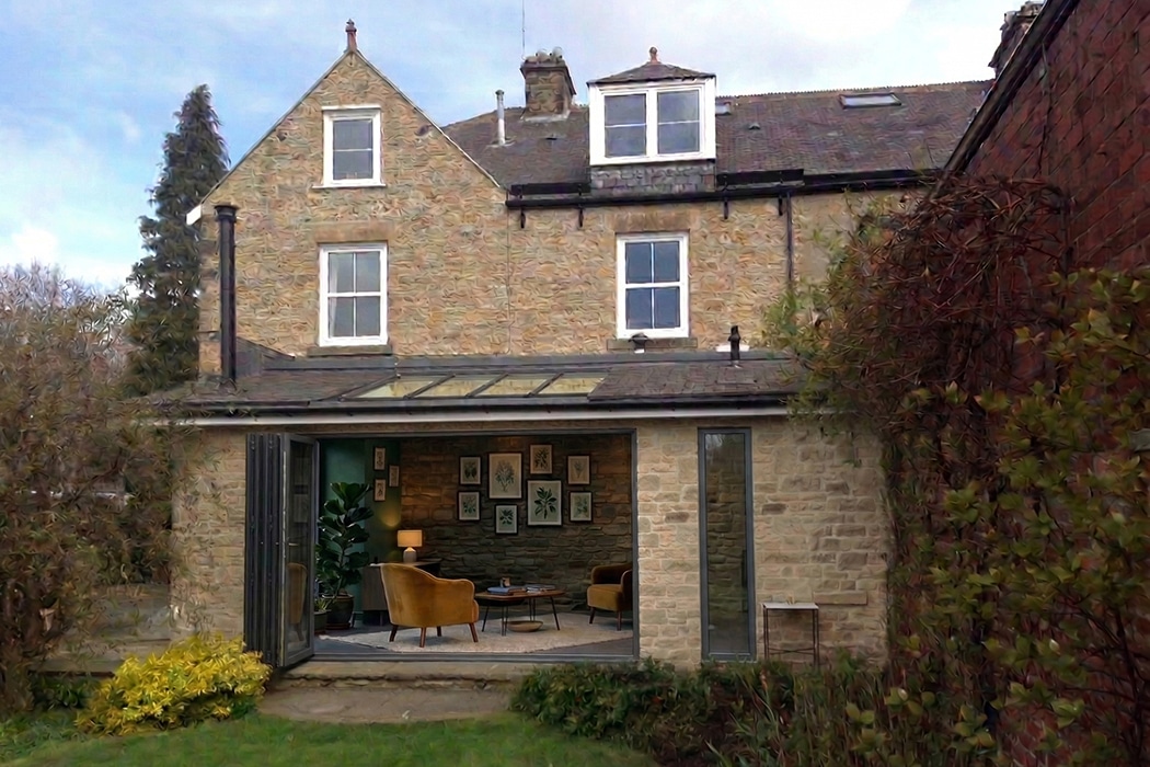 Modern stone house with inset glass roof gallery extension, featuring bifold doors opening to contemporary seating area with yellow chairs and gallery wall art.